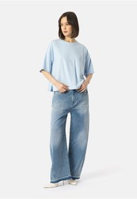 Žena stojící v lehkém modrém oversized tričku, širokých vybledlých džínách a bílých špičatých lodičkách na vysokém podpatku, na bílém pozadí.