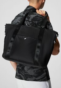 Black padded tote bag with dual webbed straps, featuring a front zip pocket and a small logo. Smooth texture and sleek design.
