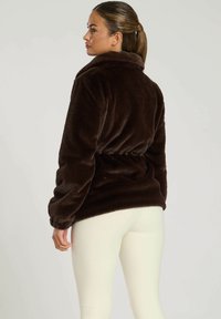 Bruine pluche fleece jack met een hoge kraag, elastische tailleband en lange mouwen, gecombineerd met crème gekleurde aansluitende leggings.