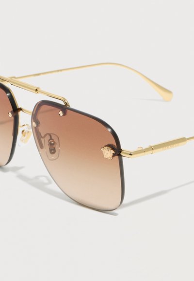 Γυαλιά ηλίου με χρυσή κορνίζα και φακούς με καφέ διαβαθμισμένο χρώμα, με έμβλημα κεφαλής Μέδουσας στη γωνία και ρυθμιζόμενα μαξιλαράκια μύτης.