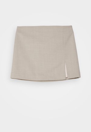 Nainen, jolla on valkoinen hihaton paita, beige minihame sivuhalkiolla, hopeinen kaulakoru ja valkoiset slip-on-tossut, seisoo harmaalla lattialla.