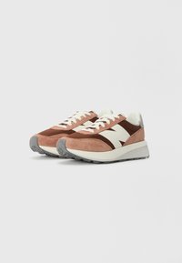 Rosa og brun ruskinnsneakers med en hvit logo, teksturerte paneler og en grå gummisåle. Har lisser og en polstret tunge.