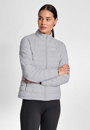 Kvinde med mørkt hår i hestehale iført en lysegrå quiltet jakke og sorte leggings, armene krydset, ser til siden.