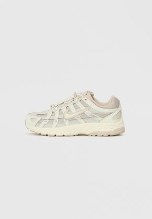 Beige og off-white Nike løbesko med lagdelt mesh og læder, snørelukning foran og chunky sål, vist fra siden på en ensfarvet baggrund.