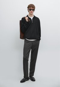 Černý pulovr s límečkem na zip, bílým tričkem pod ním, šedé kalhoty a černé mokasíny. Hnědá kožená taška a tmavé sluneční brýle.