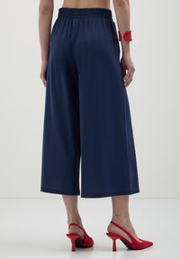 Pantalons larges bleu marine avec une taille élastique, présentant une texture lisse. Associés à des escarpins à talon rouge avec un bout pointu.