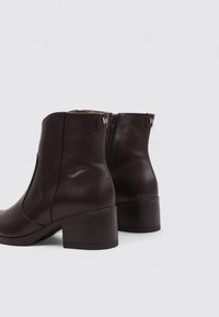 Botas de piel marrón hasta el tobillo con tacón cuadrado y cremallera lateral. Presenta acentos de logotipo en la parte posterior y una textura suave y brillante.