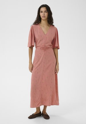 Femme debout dans une robe midi à fleurs roses avec des manches volantées, un décolleté en V, une taille froncée et des chaussures plates marron sur un fond uni.