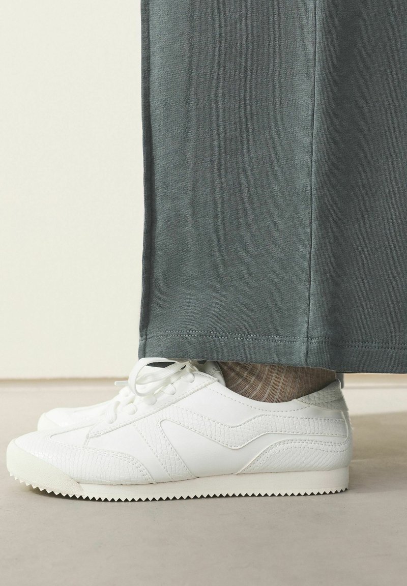 Hvide strukturerede sneakers iført med beige ribbede sokker og mørkegrønne vide bukser på et lyst gulv.