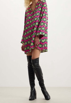 Vrouw in een langarmige jurk met een geometrisch patroon in roze en groen, gecombineerd met zwarte leren laarzen tot boven de knie, staand tegen een effen achtergrond.