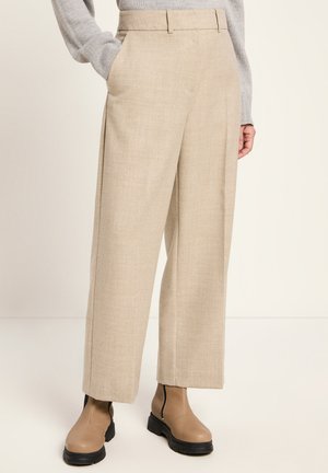 Beige Hochkante-Hosen, kombiniert mit tanfarbenen Ankle Boots und einem grauen Strickpullover, von der Taille bis zu den Füßen vor einem schlichten Hintergrund gezeigt.