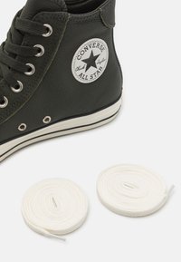 Zbliżenie ciemnozielonego sneakersa Converse All Star w stylu high-top z białą podeszwą i dwoma skręconymi białymi sznurowadłami na białym tle.