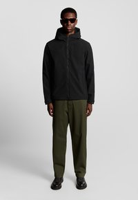Veste à capuche noire, fermeture éclair, texture douce ; associée à un pantalon vert olive et des chaussures noires à lacets avec une semelle épaisse.