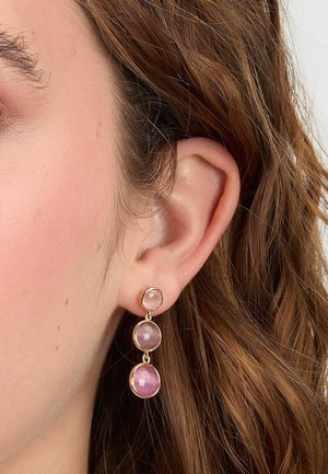 Primo piano dell'orecchio con un lungo orecchino a goccia d'oro con tre piccole pietre rosa rotonde, su uno sfondo di capelli ondulati castano chiaro.