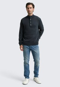 Pullover blu scuro con colletto alto, pattina con quattro bottoni, orlo a costine, abbinato a jeans azzurri chiari e sneakers bianche. Design semplice.