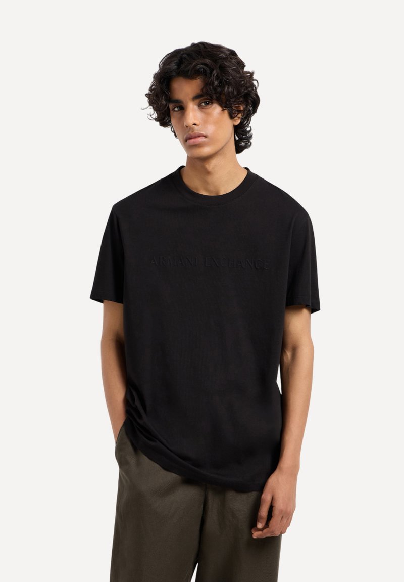 T-shirt nera a maniche corte con logo "Armani Exchange" in rilievo sulla parte anteriore, realizzata in tessuto morbido, abbinata a pantaloni larghi color oliva.
