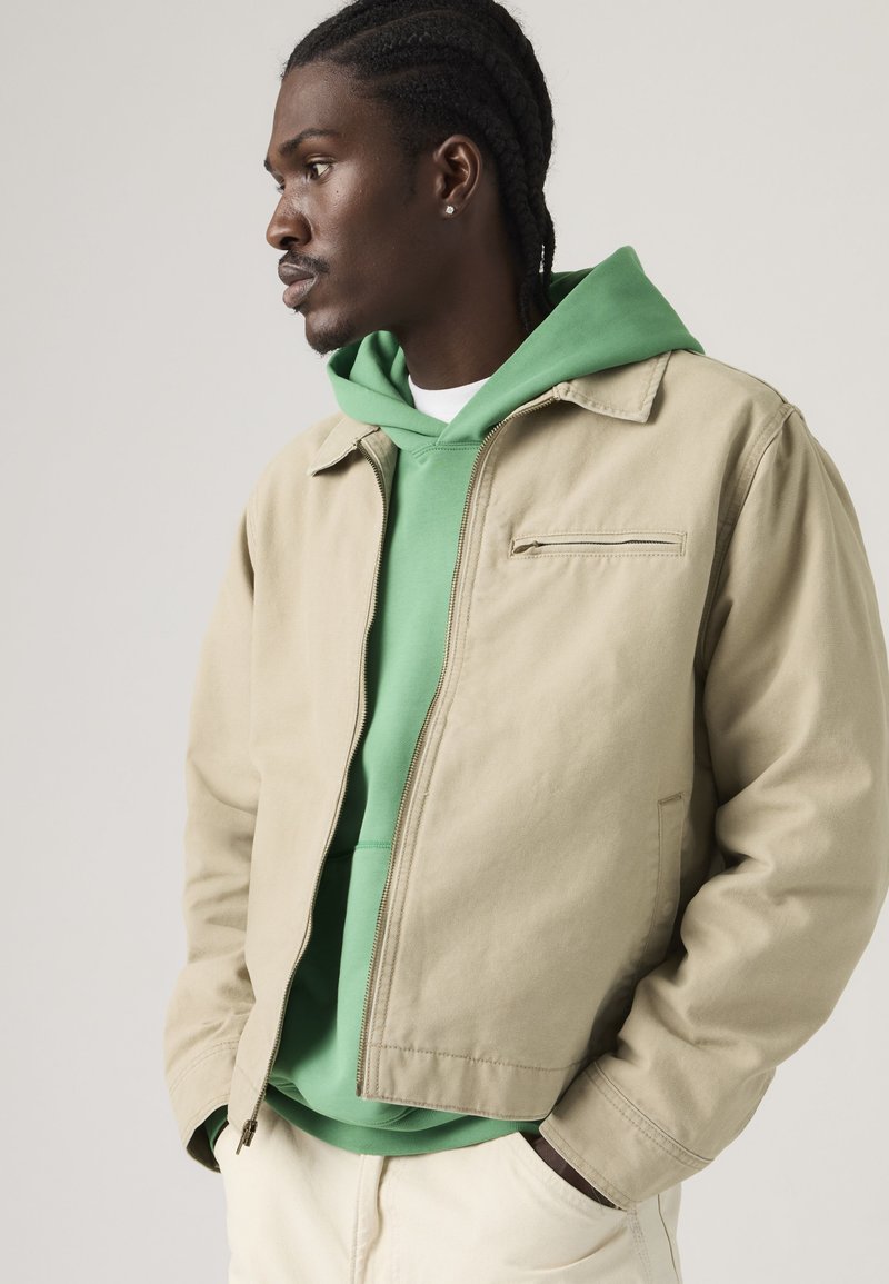 Junger Mann mit geflochtenem Haar trägt eine beige Reißverschlussjacke über einem grünen Kapuzenpullover, Hände in den Taschen, schaut nach links vor neutralem Hintergrund.