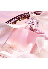 Rosa parfymeflaske med en buet design, gjennomsiktig bunn og glitrende lokk. Omgitt av hvite kronblader og en teksturert brun aksent.