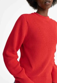 Roter gerippter Strickpullover mit Rundhalsausschnitt, strukturiertem Muster auf der Vorderseite und schmal zulaufenden Ärmeln. Der Stoff wirkt warm und dick.