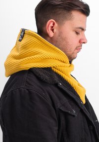 Knit yellow hooded scarf with a textured pattern, worn over a black jacket with a furry collar, featuring a logo tag at the neck.