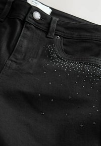 Jeans skinny negri cu talie medie, având o textură netedă și embellishments cu mici știfturi strălucitoare de-a lungul zonei buzunarului frontal.