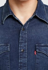 Mörkblå denimskjorta med spetsig krage, två bröstfickor och svarta knappar. Förskjuten struktur med en synlig Levi's-etikett.