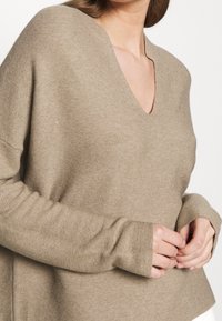 Beżowy sweter typu v-neck z długimi, dopasowanymi rękawami. Miękka dzianina i luźny krój, z bocznymi rozcięciami dla łatwości ruchu.