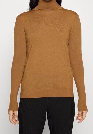 Femme portant un pull à col roulé côtelé couleur chameau et un pantalon noir texturé, vue du cou aux hanches sur un fond uni.