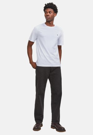 Ung mand stående iført en hvid kortærmet t-shirt, sorte bukser og mørkebrune støvler, kiggede til siden med hænderne i lommerne.