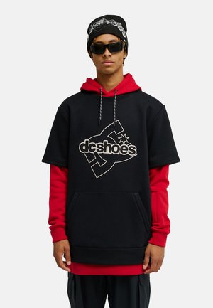 Młody mężczyzna w czarnych okularach przeciwsłonecznych, czarnej czapce i warstwowych czerwono-czarnych bluzach z logo "dc shoes" z przodu.