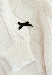 Weißer Strickpullover mit einem strukturierten Muster, Kabeldesign und einem schwarzen Schleifenakzent. Gerippte Ärmelbündchen mit einem weißen Rüschendetail.