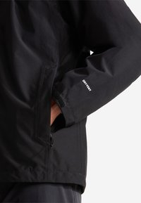 Personne portant une veste noire avec le texte « DRY VENT » sur la manche, main partiellement dans la poche avant.