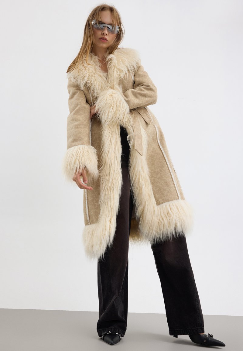 Manteau en fausse fourrure beige à manches longues, avec un col et des poignets en peluche, assorti à un pantalon noir à jambes larges et des chaussures noires à bout pointu.