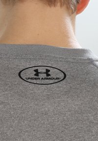 Grå t-shirt med kort ärm som har en svart Under Armour-logga i oval design placerad på nackområdet baksidan. Slät tygtextur.
