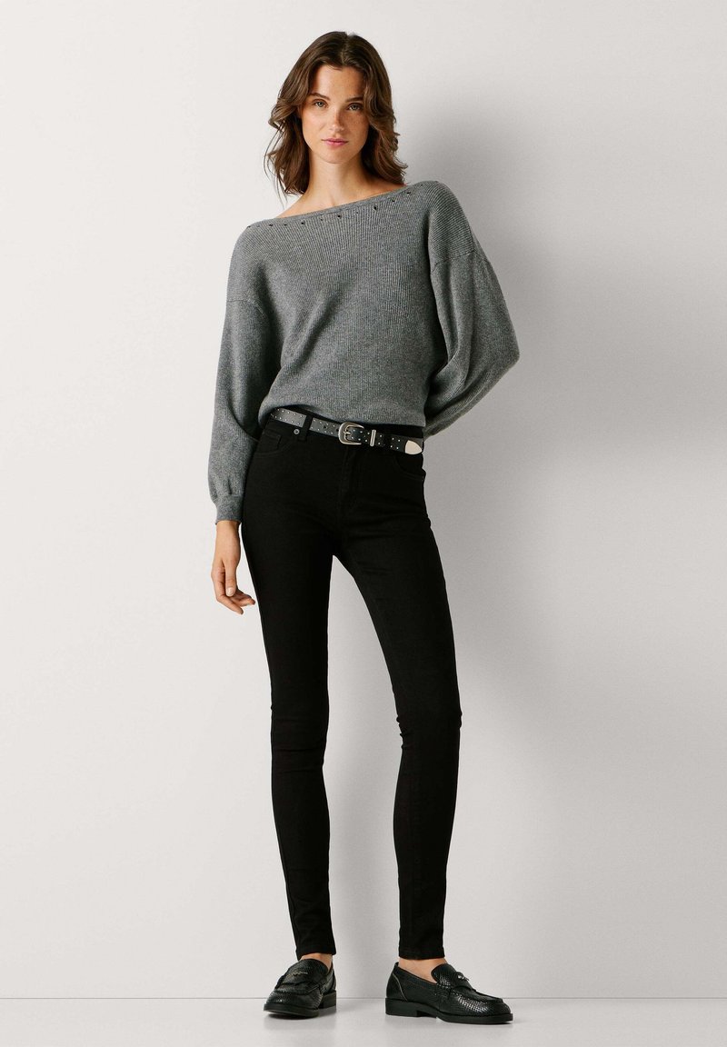 Suéter gris texturizado con hombros caídos, jeans negros ajustados y zapatos negros con cordones, con un cinturón brillante con detalles de hebilla.