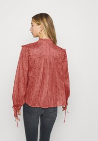 Blusa roja con estampado y hombros con volantes y puños con lazo. Tejido ligero con pliegues verticales y pequeños motivos en toda la prenda. Vista trasera mostrada.