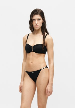 Nuori nainen seisoo musta bikini päällä, jossa on ohuet olkaimet ja valkoiset yksityiskohdat, yksinkertaisen valkoisen taustan edessä.