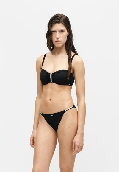 Jeune femme debout, portant un bikini noir avec de fines bretelles et des accents blancs sur un fond blanc uni.