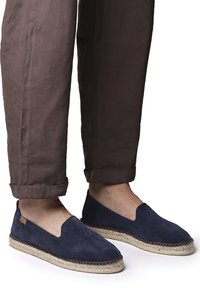 Námornícky modré semišové espadrilky s tkanou jútovou podrážkou, s designem na obutí a bočními výřezem. Hnědé kalhoty s manžetou jsou částečně viditelné.