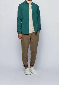 Teal button-up shirt layered over a beige turtleneck, paired with brown tapered pants and white lace-up shoes.