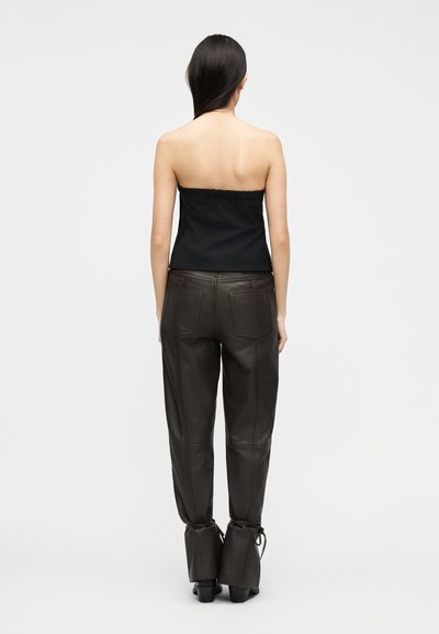 Γυναίκα με μακριά σκουρόχρωμα μαλλιά που φοράει μαύρο τοπ χωρίς τιράντες και σκούρο καφέ δερμάτινο παντελόνι με πίσω τσέπες, στέκεται μπροστά σε ένα απλό φόντο.