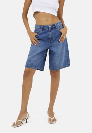 Kobieta w luźnych dżinsowych szortach z wysokim stanem i białych sandałach na obcasie, z rękami w kieszeniach, ubrana w krótki biały top.