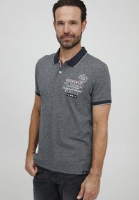 Sivý polo tričko s kontrastným námorníckym golierom. Obsahuje grafický potlač na hrudi a krátke rukávy. Slim fit s bočnými rozparky.