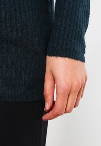 Dzianinowy sweter w ciemnym odcieniu turkusu o prążkowanej teksturze, z dopasowanymi długimi rękawami i lekko postrzępionym dołem. Ręka oparta na czarnym materiale.
