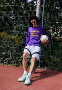 Lila långärmad tröja med texten "LOS ANGELES LAKERS BASKETBALL" i gult, vita shorts med svarta ränder och ljusgröna sportskor. Håller en basketboll.