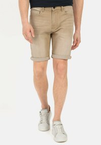 Shorts en denim beige avec ourlets roulés, design à cinq poches et fermeture à bouton à l'avant, associés à des baskets blanches sur un fond uni.