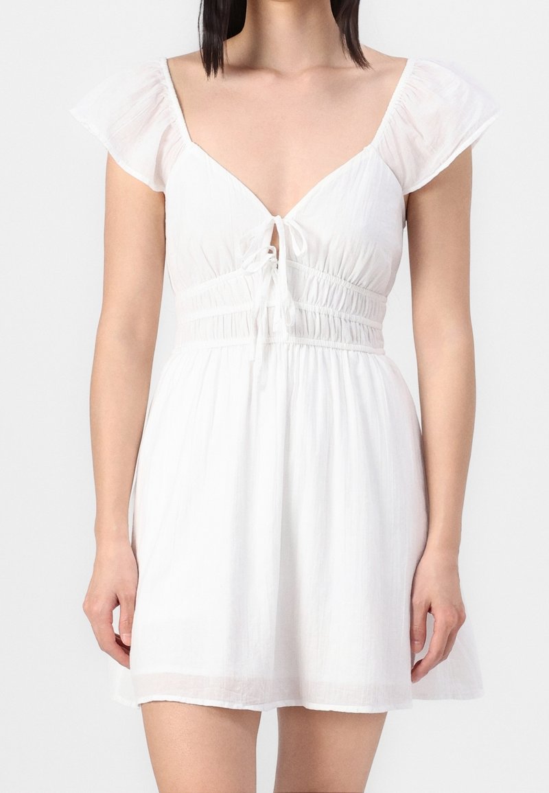 Vit kort klänning med fladderärmar, djup V-ringning och åtsittande midja. Tillverkad av lätt tyg med en mjuk textur och justerbar snöre.