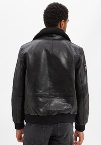 Zwarte leren jas met een zachte zwarte schapenvacht kraag, geribde manchetten en zoom, en een patchdetail op de rechter schouder.