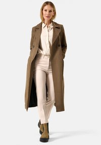 Bruine wollen blend lange jas met een dubbelgeknopte sluiting, gedragen over een lichtblouse en lichtroze pantalon. Tan enkellaarsjes met zwarte accenten.