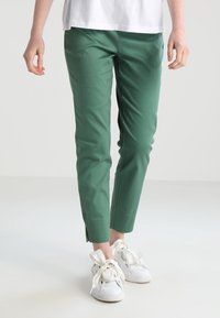 Pantalons verts ajustés en tissu lisse, avec un design épuré, de subtils fentes sur l'ourlet, associés à des baskets blanches.